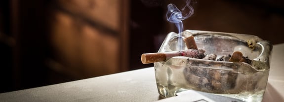 A lit cigarette resting on the edge of a glass ash tray filled with used cigarette butts.