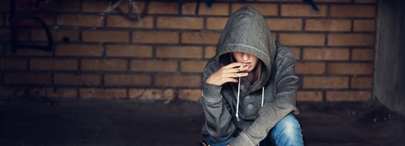 Young adult female wearing a hooded sweatshirt, smoking a cigarette and looking pensive.