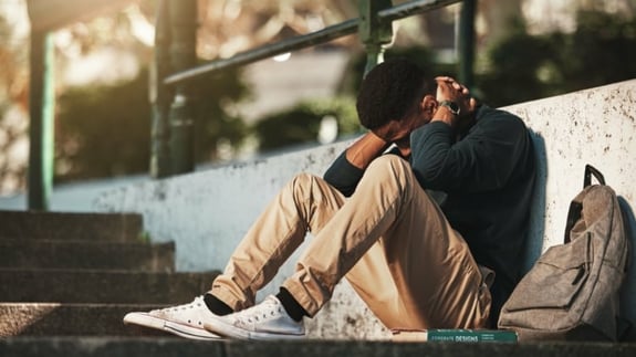 Young adult male sitting outside on the ground. His knees are bent and his hands are clasped behind his neck with his elbows on his knees.  He looks upset