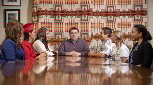 A group of diverse aduts sitting at a table having a discussion. A Native American tapistry hangs on the wall in the background.