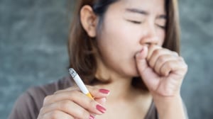 Young adule female holding a lit cigarette and coughing into her other hand.